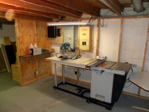 View of table saw and electrical panels [Verizon FiOS (l), circuit breaker panel (c), Guardian Generac Circuit panel (r)]. Newly installed 220v (white) outlet for table saw is visible just above center of saw. Small stud wall on left will be removed.